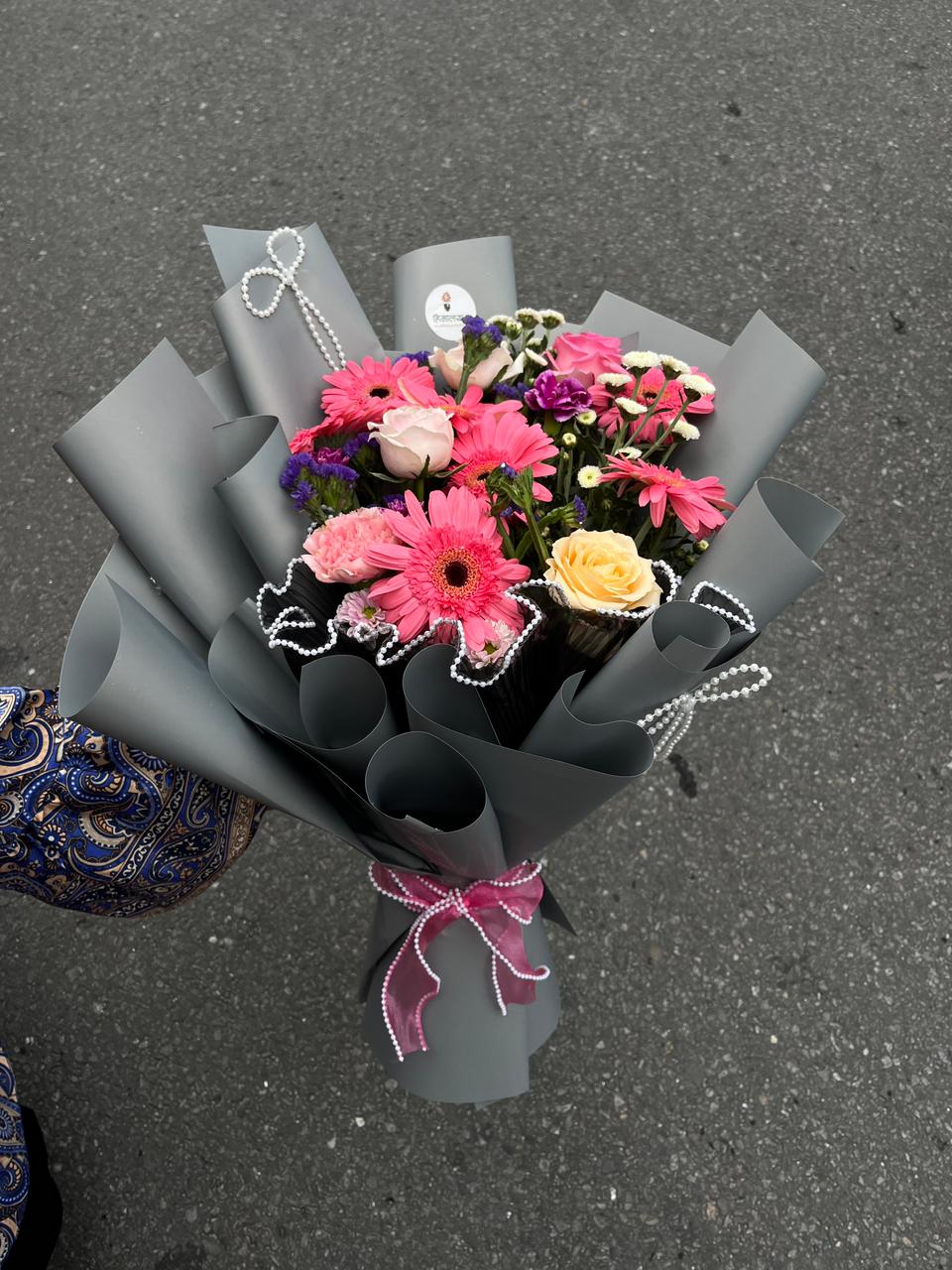 A 15-stem bouquet with roses, gerbera daisies, and seasonal filler flowers in gray wrapping paper.